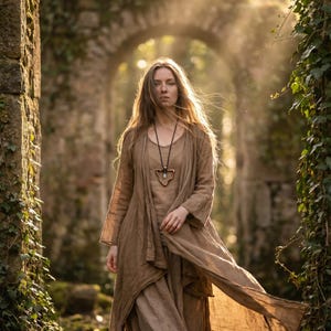 May include: A woman in a long, flowing brown dress and matching duster coat stands in a stone archway. She wears a necklace with a pendant. The scene is set in a sunlit forest with ivy-covered stone walls.