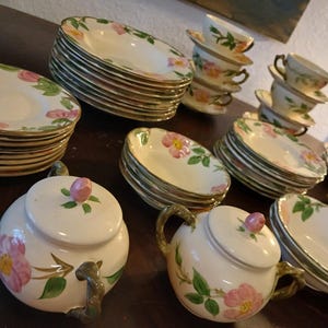 May include: A vintage porcelain tea set featuring plates, saucers, cups, and serving pieces. The set is decorated with pink rose floral patterns and green leaves. The plates and saucers have a scalloped edge.