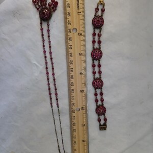 Puede incluir: Un conjunto de joyas vintage con collar y pulsera. El collar tiene un colgante con cuentas colgantes y una cadena larga. La pulsera tiene tres eslabones redondos y ornamentados. Ambas piezas son de color rojo oscuro y miden unos 25 cm de largo.