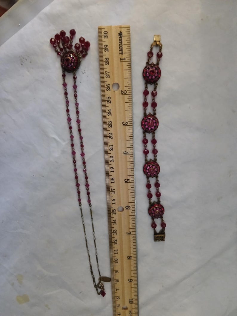 Puede incluir: Un conjunto de joyas vintage con collar y pulsera. El collar tiene un colgante con cuentas colgantes y una cadena larga. La pulsera tiene tres eslabones redondos y ornamentados. Ambas piezas son de color rojo oscuro y miden unos 25 cm de largo.