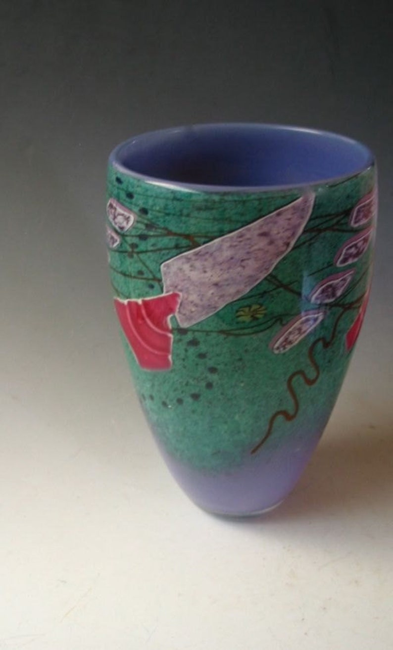 May include: A tapered glass vase featuring a vibrant green body with a speckled texture, accented by abstract purple and red shapes, thin dark brown lines, and a solid light purple rim that transitions into a light purple base at the bottom.