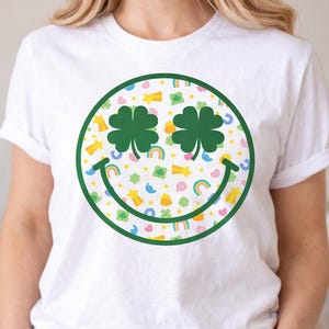 May include: White t-shirt featuring a large green-outlined smiley face with shamrock eyes and a smiling mouth. The face is filled with a pattern of rainbows, stars, and other St. Patrick's Day themed graphics.