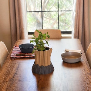 May include: A cylindrical planter with a vertical wood-grain pattern sits on a dark gray, rock-textured base. A green herb plant grows inside the planter. The planter is placed on a wooden dining table with two bowls and a cloth napkin.