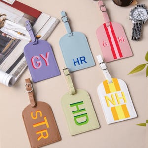 May include: Six rectangular luggage tags with adjustable buckle straps. Colors include purple with GY, light blue with HR, pink with CM, brown with STR, light green with HD, and white with NH. Each tag features bold, colorful lettering.