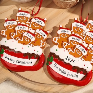 May include: Two red mug-shaped Christmas ornaments featuring gingerbread figures wearing Santa hats. The mugs have white rims with green and red dots, and red ribbons for hanging. Names are printed on the hats, with Merry Christmas or 2026 and The Smith family on the mugs.