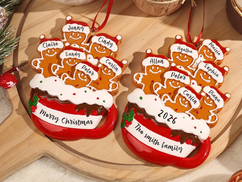 May include: Two red mug-shaped Christmas ornaments featuring gingerbread figures wearing Santa hats. The mugs have white rims with green and red dots, and red ribbons for hanging. Names are printed on the hats, with Merry Christmas or 2026 and The Smith family on the mugs.