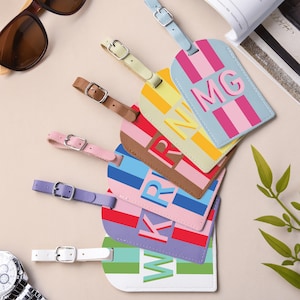 May include: A collection of six rectangular luggage tags with rounded edges, each featuring vertical stripes in various colors and a large monogram letter or letters printed in the center. Each tag includes an adjustable strap with a metal buckle.