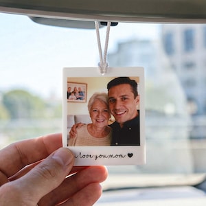May include: A white, rectangular car air freshener with a photo of a smiling woman and man. The text "i love you mom" is printed at the bottom, with a small heart symbol. The air freshener hangs from a white string.