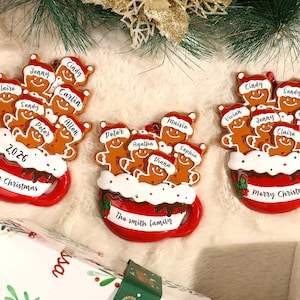 May include: Three red mug-shaped Christmas ornaments featuring brown gingerbread figures wearing Santa hats. Each mug has white frosting, red and green holly accents, and text including Merry Christmas, 2026, or The Smith family.