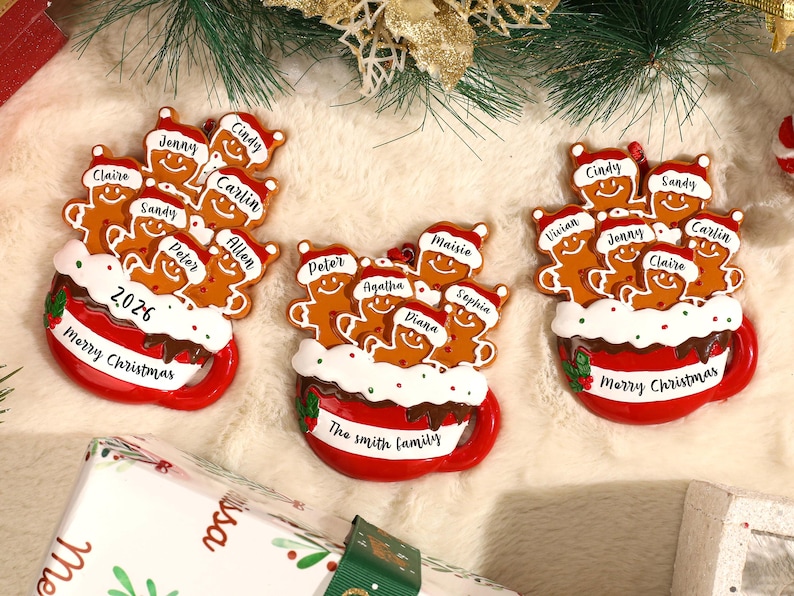 May include: Three red mug-shaped Christmas ornaments featuring brown gingerbread figures wearing Santa hats. Each mug has white frosting, red and green holly accents, and text including Merry Christmas, 2026, or The Smith family.