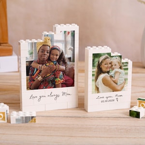 Könnte beinhalten: Zwei weiße Fotorahmen im Ziegelstil mit Familienfotos. Ein Rahmen trägt den Text "Love you always, Mom". Der andere Rahmen trägt den Text "Love you, Mom 05.10.2026". Zusätzliche weiße und gelbe Ziegelsteine sind verstreut.