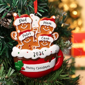 May include: A Christmas tree ornament shaped like a red mug filled with four gingerbread figures wearing Santa hats. The figures are labeled Dad, Mom, Peter, and Coco. The mug features the text 2026 and Merry Christmas with green holly leaves.