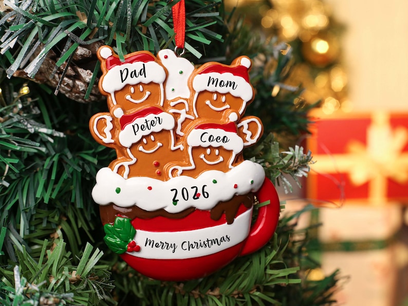 May include: A Christmas tree ornament shaped like a red mug filled with four gingerbread figures wearing Santa hats. The figures are labeled Dad, Mom, Peter, and Coco. The mug features the text 2026 and Merry Christmas with green holly leaves.