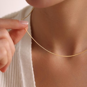 May include: A delicate gold-colored necklace is held near a person's neck. The necklace is a thin, curved line, suggesting a minimalist design. The person is wearing a white sweater, and the background is a soft, neutral tone.