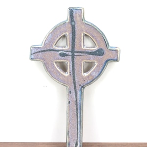 May include: A ceramic Celtic cross featuring a mottled lavender and blue glaze with a dark, thin, vertical charcoal-colored line running down the center. The cross has a circular ring intersecting the arms and a smooth, glossy, reflective finish.