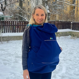 May include: A woman wearing a navy blue baby carrier with a large front pocket and the word "Boba" in green. The carrier is worn over a gray sweater and black pants. The background is a snowy outdoor scene.