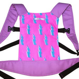 May include: A purple baby carrier with adjustable straps and a pink panel featuring a white and blue lightning bolt pattern. The carrier has a label that reads "Bo-Bo".