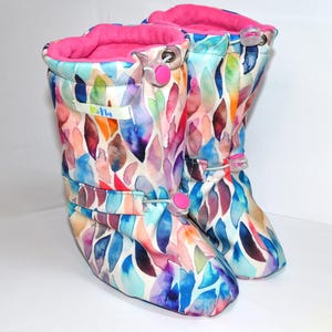 May include: A pair of colorful baby boots with a watercolor leaf pattern in shades of blue, purple, orange, and green. The boots have a pink fleece lining and pink toggle closures. The boots have the word "Rella" printed on the side.