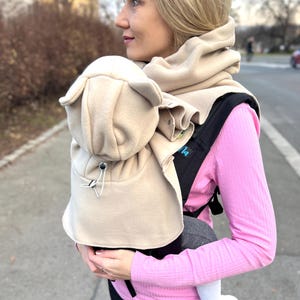 May include: A beige baby carrier with a matching hood and scarf. The carrier is black and is being worn by a person wearing a pink long-sleeved shirt and black pants. The baby is wearing a white sock.