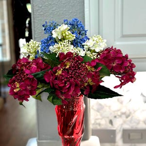 Puede incluir: Un ramo de flores artificiales con hortensias de color magenta intenso, azul brillante y blanco con hojas verdes, dispuesto en un jarrón de cristal rojo translúcido con un patrón texturizado en espiral y una base hexagonal transparente.