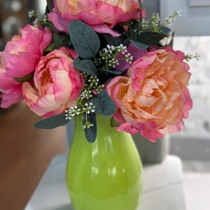 Può includere: Una vivace composizione di peonie artificiali in un vaso verde brillante. I fiori presentano sfumature di rosa e pesca, completate da foglie verde scuro e piccoli accenti bianchi. Il vaso ha una finitura lucida.