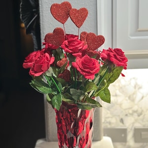 May include: A red glass vase filled with red roses and glittery heart decorations. The vase has a textured surface and is filled with artificial roses and heart-shaped picks, creating a romantic floral arrangement.