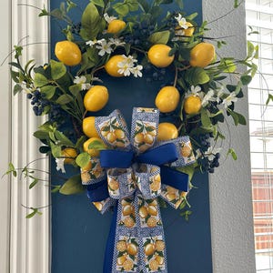 May include: A decorative wreath featuring bright yellow lemons, dark blue berries, and white flowers. The wreath is adorned with green leaves and a large bow with a lemon and blue pattern. The wreath is hanging on a dark blue wall.