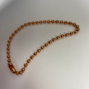 Può includere: Una collana a catena a sfere color oro con chiusura. La catena è composta da piccole sfere distanziate uniformemente. La chiusura è in tinta oro. La collana è esposta su una superficie bianca.