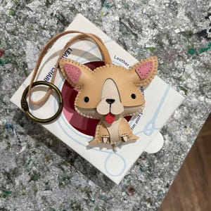 May include: A tan leather keychain shaped like a dog with pink ears and a red tongue. The keychain is attached to a brown leather strap and a metal ring.