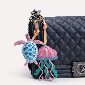 Puede incluir: Un bolso de mano acolchado negro con una correa de cadena de cuero y un charm con una medusa rosa y una tortuga azul y rosa.