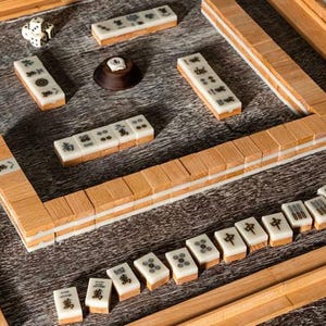 Puede incluir: Un juego de Mahjong de madera con fichas de color marfil con s&iacute;mbolos y caracteres negros. El juego incluye dados, un soporte de fichas de madera y un tablero de juego de madera. Las fichas est&aacute;n dispuestas sobre una superficie de madera.