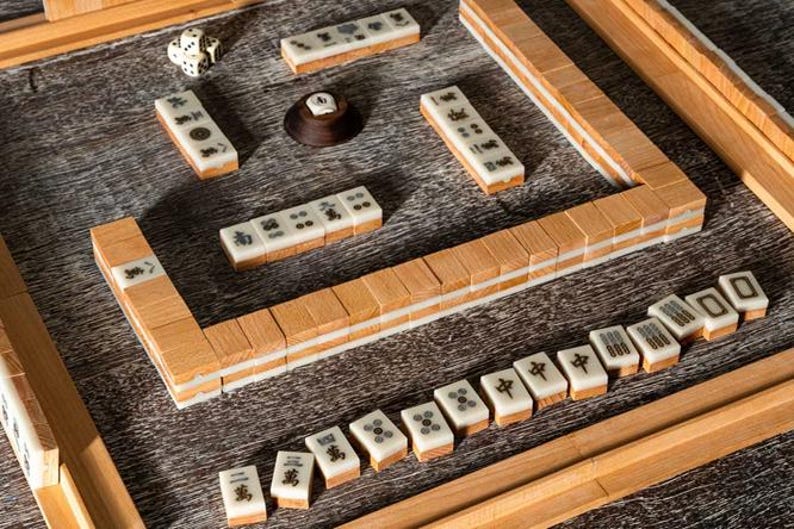 Puede incluir: Un juego de Mahjong de madera con fichas de color marfil con s&iacute;mbolos y caracteres negros. El juego incluye dados, un soporte de fichas de madera y un tablero de juego de madera. Las fichas est&aacute;n dispuestas sobre una superficie de madera.