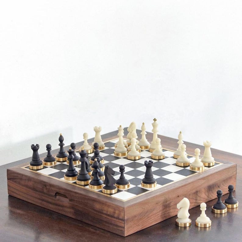 May include: A complete chess set with black and cream-colored pieces, each with a gold-colored base, arranged on a checkered board. The board is set within a wooden box with a dark finish. The chess pieces are ready for play.