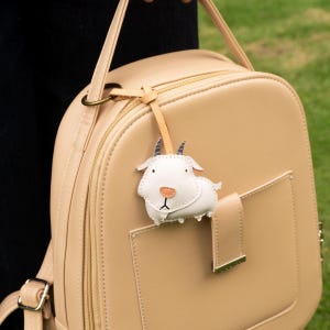 May include: A beige backpack with a small, white goat-shaped leather tag attached. The tag has black horns and a peach-colored nose. The backpack has a front pocket and a gold-colored clasp.