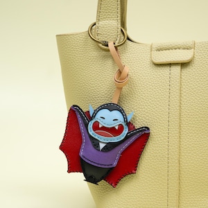 Könnte beinhalten: Ein lederner Taschenanhänger in Form eines Comic-Vampirs. Der Anhänger hat ein blaues Gesicht, einen roten Umhang, eine lila Weste und schwarze Details. Er ist an einem beigefarbenen Lederriemen und einem goldfarbenen Metallring befestigt und hängt an einer beigen Handtasche.