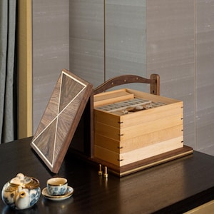 Puede incluir: Una caja de joyer&iacute;a de madera con tapa con bisagras, que presenta un dise&ntilde;o geom&eacute;trico con detalles en colores claros. La caja est&aacute; abierta, revelando m&uacute;ltiples compartimentos. Una peque&ntilde;a tetera y una taza de t&eacute; est&aacute;n sobre la mesa frente a la caja.