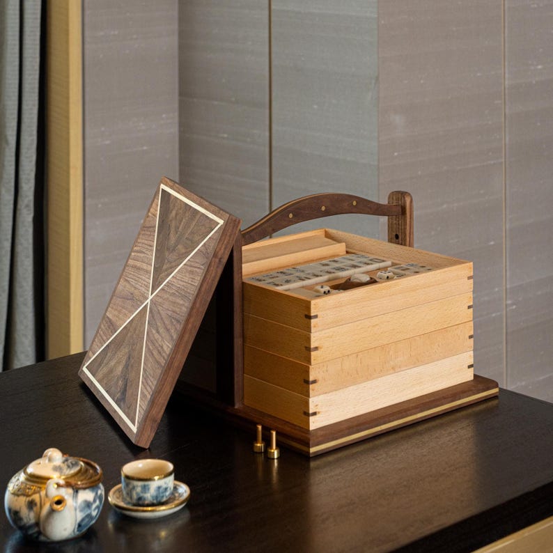 Puede incluir: Una caja de joyer&iacute;a de madera con tapa con bisagras, que presenta un dise&ntilde;o geom&eacute;trico con detalles en colores claros. La caja est&aacute; abierta, revelando m&uacute;ltiples compartimentos. Una peque&ntilde;a tetera y una taza de t&eacute; est&aacute;n sobre la mesa frente a la caja.