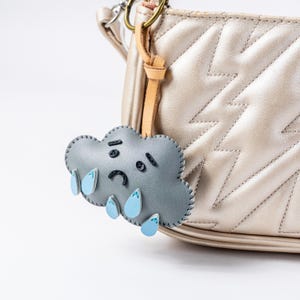 May include: A gray leather cloud-shaped keychain with a sad face and blue raindrops hanging from it. The keychain is attached to a gold-colored chain and is hanging from a light brown leather bag.