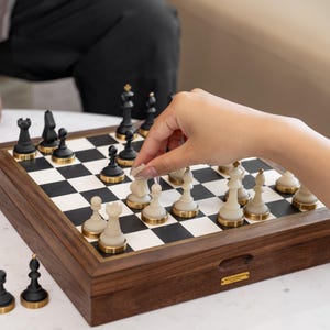 May include: A wooden chess set with black and cream-colored pieces. The chess board is set up with pieces in play. The pieces have gold-colored accents. The board is in a wooden box with a gold-colored label.