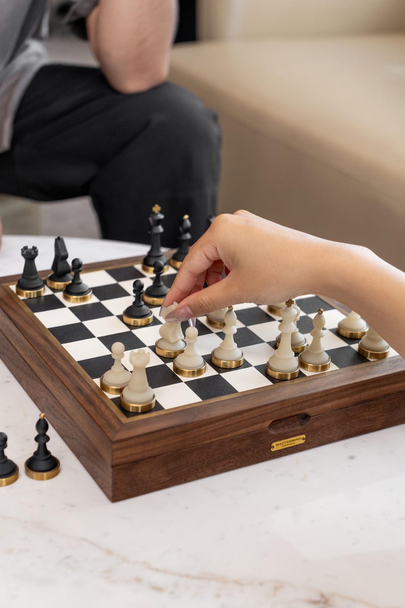 May include: A wooden chess set with black and cream-colored pieces. The chess board is set up with pieces in play. The pieces have gold-colored accents. The board is in a wooden box with a gold-colored label.