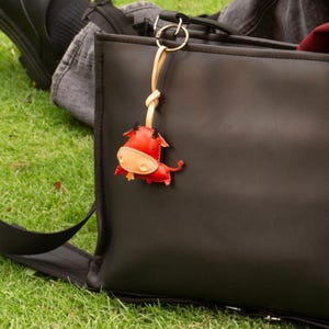 May include: A red and tan leather bull keychain with black horns and a gold-colored metal clasp. The keychain is attached to a black bag, with green grass in the background.