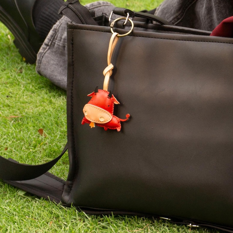 May include: A red and tan leather bull keychain with black horns and a gold-colored metal clasp. The keychain is attached to a black bag, with green grass in the background.