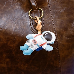 Puede incluir: Un bolso tote de cuero marrón con un pequeño dije de astronauta. El dije es blanco, azul y rojo, con una visera negra. El dije está sujeto al asa del bolso con un anillo dorado y una correa de cuero color beige.