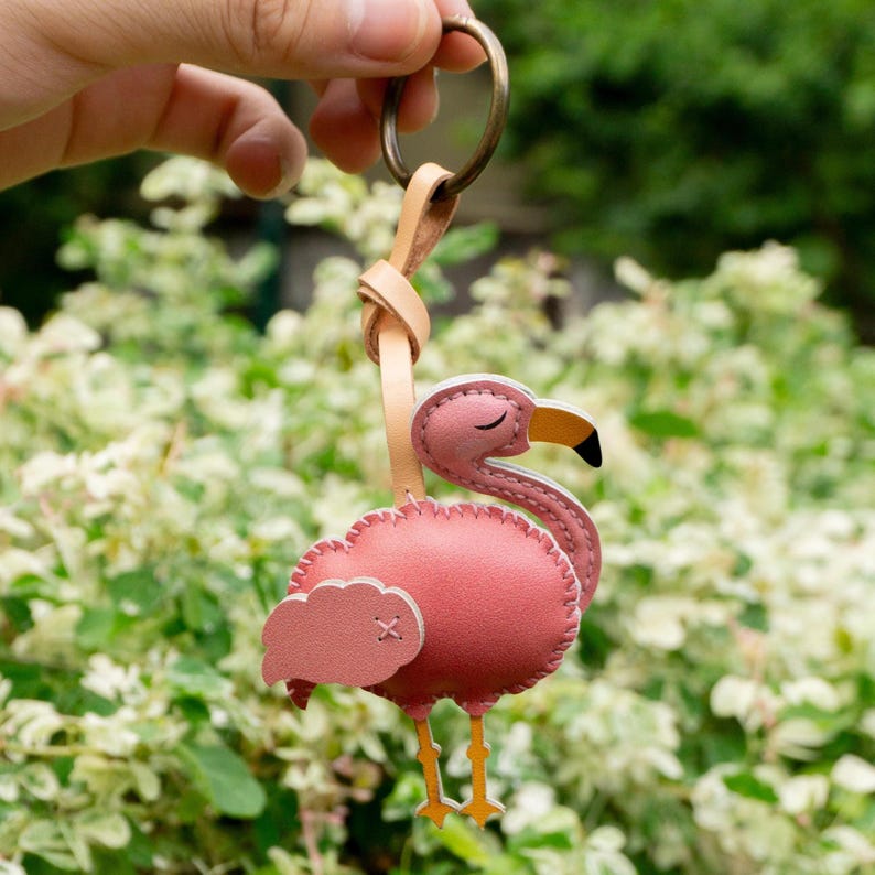 May include: A pink flamingo keychain with a leather strap and metal ring. The flamingo has a closed eye, a yellow beak with a black tip, and yellow legs. The keychain is held up by a hand against a blurred green background.