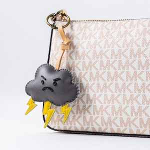Könnte beinhalten: Ein grauer Leder-Wolken-Schlüsselanhänger mit einem gelben Blitz-Design. Der Schlüsselanhänger ist an einem braunen Lederband und einem goldenen Metallring befestigt.