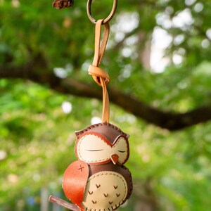 May include: A brown and tan leather owl keychain with a closed-eye design, hanging from a metal ring and leather strap. The owl is perched on a small branch, with a red-orange wing. The background is a blurred green.