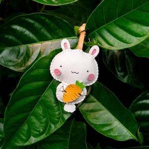 May include: A white bunny-shaped keychain with pink accents and a stitched outline, holding an orange carrot. The keychain has a leather strap and is set against a backdrop of lush green leaves.