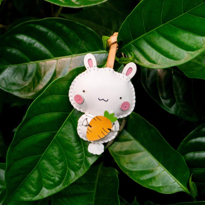 May include: A white bunny-shaped keychain with pink accents and a stitched outline, holding an orange carrot. The keychain has a leather strap and is set against a backdrop of lush green leaves.