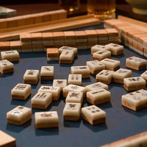 Puede incluir: Un conjunto de fichas de Mahjong de color marfil y marr&oacute;n dispersas sobre una superficie azul. Las fichas tienen varios s&iacute;mbolos y caracteres. Tambi&eacute;n se ven estanter&iacute;as de madera y otras piezas del juego, lo que sugiere un juego en curso.