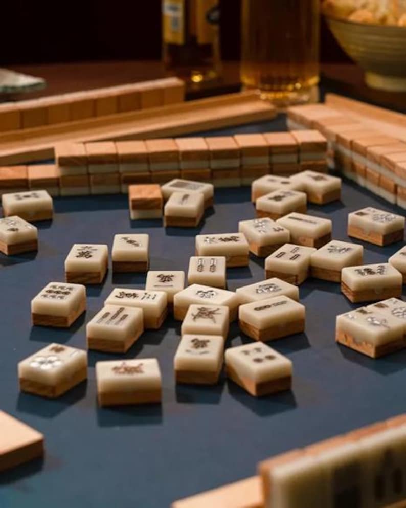 Puede incluir: Un conjunto de fichas de Mahjong de color marfil y marr&oacute;n dispersas sobre una superficie azul. Las fichas tienen varios s&iacute;mbolos y caracteres. Tambi&eacute;n se ven estanter&iacute;as de madera y otras piezas del juego, lo que sugiere un juego en curso.